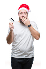 Sticker - Young caucasian man wearing christmas hat holding credit card over isolated background cover mouth with hand shocked with shame for mistake, expression of fear, scared in silence, secret concept