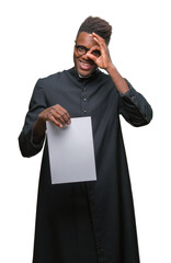 Canvas Print - Young african american priest man over isolated background holding blank paper with happy face smiling doing ok sign with hand on eye looking through fingers