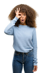 Poster - African american woman wearing a sweater doing ok gesture shocked with surprised face, eye looking through fingers. Unbelieving expression.