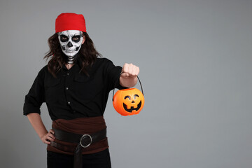 Poster - Man in scary pirate costume with skull makeup and pumpkin bucket on light grey background, space for text. Halloween celebration