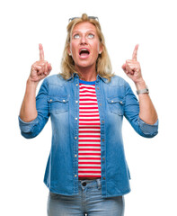 Poster - Middle age blonde woman over isolated background amazed and surprised looking up and pointing with fingers and raised arms.