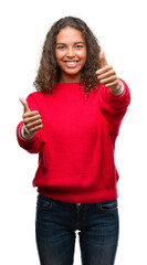 Wall Mural - Young hispanic woman wearing red sweater approving doing positive gesture with hand, thumbs up smiling and happy for success. Looking at the camera, winner gesture.