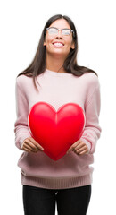 Poster - Young hispanic woman holding heart in love with a happy face standing and smiling with a confident smile showing teeth