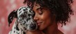 Close-up portrait of overjoyed African American young woman hugging and kissing her Dalmatian dog. Happy beautiful dark skinned girl with devoted pet. Isolated on pink background.