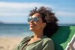 Happy young black woman relaxing on deck chair at beach wearing spectacles. Smiling african american girl with sunglasses enjoy vacation. Carefree happy young woman sunbathing at sea with copy space.