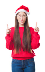 Sticker - Young Chinese woman over isolated background wearing christmas hat amazed and surprised looking up and pointing with fingers and raised arms.