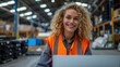 © LoLArt - Happy smiling curly blonde woman wearing a vest with reflective stripe in light warehouse worker sitting at desk with her laptop looking at camera with toothy smile at logistic centre