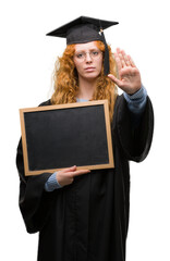 Wall Mural - Young redhead student woman wearing graduated uniform holding blackboard with open hand doing stop sign with serious and confident expression, defense gesture