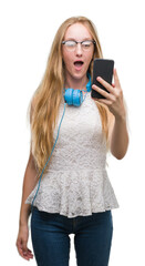 Sticker - Blonde teenager woman holding smartphone and wearing headphones scared in shock with a surprise face, afraid and excited with fear expression