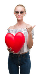 Sticker - Blonde teenager woman holding red heart pointing and showing with thumb up to the side with happy face smiling