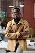 © AnnaStills - Young african american office worker in smart casual outfit confidently posing against his workplace