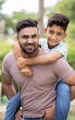 Vertical shot of joyful indian father piggy back his son by looking at camera at park - concept of father love, playful and relationship