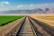 Train tracks leading through green fields to mountains under blue sky