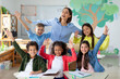 © Home-stock - Excited diverse schoolchildren raising arms and smiling, sitting in classroom with their happy female teacher