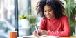 © Attasit - Happy pretty American African woman fashion designer writing ideas for small business in notebook