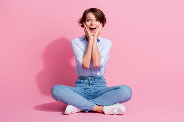 Full size photo of astonished lovely girl sit floor crossed legs hands touch cheeks isolated on pink color background