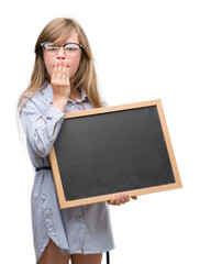 Sticker - Young blonde child holding blackboard cover mouth with hand shocked with shame for mistake, expression of fear, scared in silence, secret concept