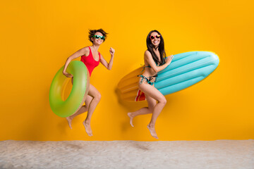 Sticker - Full length photo of excited funky ladies wear bikini jumping high running swimming buoys isolated yellow color background