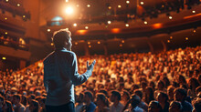 Keynote Speaker Giving A Lecture To A Large Audience In A Grand Auditorium The Speaker Is Confidently Addressing The Crowd, Gesturing With Their Hand, Under The Spotlight On Stage