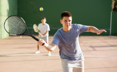 Wall Mural - Emotional athletic young guy playing frontenis on open court on summer day, hitting ball with strung tennis racquet to score to opposing team