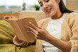 © Chanakon - Asian beautiful woman sitting reading book on cozey couch sofa in living room with sun light morning. People female stay home part time relax at home.