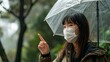 Young Asian woman caught virus during wet autumn day, wears medical mask not to spread infection points fore finger on blank space right poses under umbrella protects herself from being caught in rain