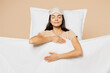 Young calm pregnant Latin woman wears pyjamas jam sleep eye mask rest relax at home under duvet hold hands on belly isolated on plain pastel light beige background studio. Good mood night nap concept.