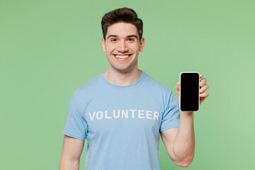 Sticker - Young man wearing blue t-shirt white title volunteer hold use mobile cell phone with blank screen isolated on plain pastel green background. Voluntary free work assistance help charity grace concept.