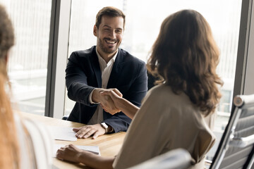 Wall Mural - Positive millennial business partners shaking hands on corporate meeting. Confident businessman giving greeting, handshake to female colleague over table, smiling, laughing