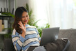 © Prathankarnpap - Beautiful young woman in sweater sitting on sofa at home and having video call via laptop