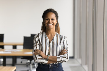 Positive beautiful young Black business woman posing in office with hands folded, looking at camera with toothy smile. Happy female entrepreneur, professional, worker girl head shot portrait