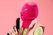 Beautiful young stylish woman in balaclava with gun on pink background