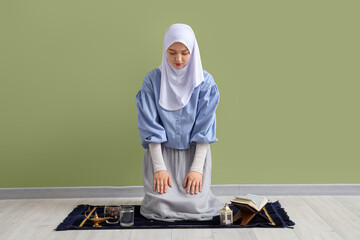 Wall Mural - Young Muslim woman praying on mat near green wall. Ramadan celebration