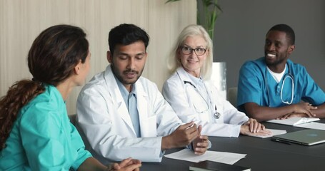 Wall Mural - Young Indian doctor man in white coat talking to hospital job colleagues at meeting table, offering ideas for discussion, teamwork, discussing patients cases, diagnosis with diverse team