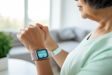 Over the shoulder view of senior Asian woman resting after exercising at home, looking at her smart watch, using fitness tracker app and measuring pulse. Maintaining healthy fitness habits. Elderly we