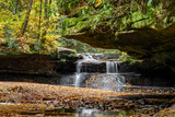 Fototapeta  - Creation Falls, a waterfall in the Red River Gorge region of Kentucky, cascading over a rocks covered with colorful autumn leaves, is viewed downstream from under an overhanging rock ledge.