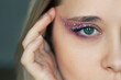 Close up of a young caucasian blonde woman's green eye with purple and pink eye shadow and glitter on the eyelid. Festive makeup. Party, holiday makeover