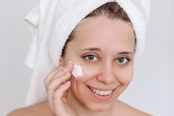 Wall Mural - Close up of a young caucasian attractive smiling woman with towel on her head after shower applying a moisturizing cream on the skin of face with her hand on a light background. Cosmetology and beauty