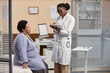 © Seventyfour - Cheerful African American female patient sitting on examination table while young woman doctor filling in medical card standing beside in modern clinic office