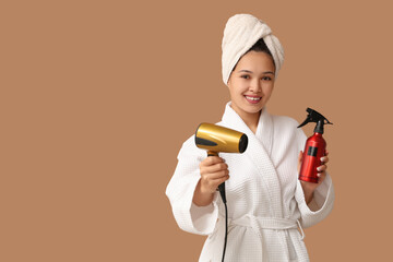 Sticker - Beautiful young Asian woman in bathrobe with hair dryer and spray on brown background