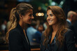 © sommersby - Two women smiling at each other. One has blonde hair and black blazer while other has brunette hair and black shirt. They are standing in crowded room with various people. Blurred background with bar