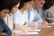 © fizkes - Focused multiracial millennial students sit in row at desk busy answering questions writing test or exam, concentrated multiethnic young people handwriting making notes summarize lecture in classroom