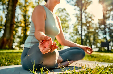 Wall Mural - Yoga training outdoors. Crop of relaxed aged woman holding hands in mudra gesture while meditating on yoga mat in lotus pose. Healthy fit pensioner gaining harmony and zen under morning sun at park.