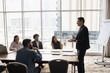 © fizkes - Confident young team leader man talking to multiethnic colleagues on corporate meeting in modern urban office interior, speaking at conference table with large window and city view