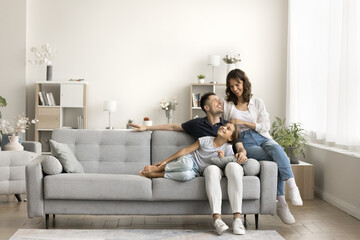 Happy couple of young parents and little daughter kid sitting and relaxing on sofa, resting on comfortable couch in modern cozy home interior, talking, laughing, smiling. Full length wide shot