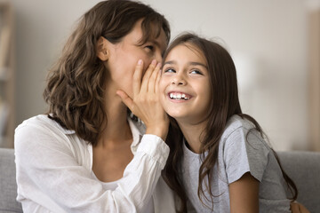  Happy playful mother telling secret to cute pretty girl kid, whispering good funny news at ear, enjoying family bonding, communication, motherhood, leisure with little daughter at home