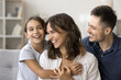 © fizkes - Carefree cute little girl kid hugging positive mom and dad on couch, smiling, laughing. Happy couple of parents playing with child on sofa, enjoying care, family leisure time at home