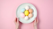 Two hands holding plate filled with colored Easter eggs on a soft pink background. Top view. Suitable for Easter event promotions and seasonal decor themes.
