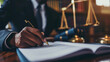 Close-up of a person's hands signing a document, with a pen in one hand with a blurred background featuring a scale of justice, symbolizing a legal setting or the act of signing important documents.