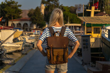 Young Female Tourist With A Craft Backpack Stands On The Pier Near Yachts And Boats At Sunset, Back Photo, Marmaris, Turkey. Concept Of Travel And Tourism